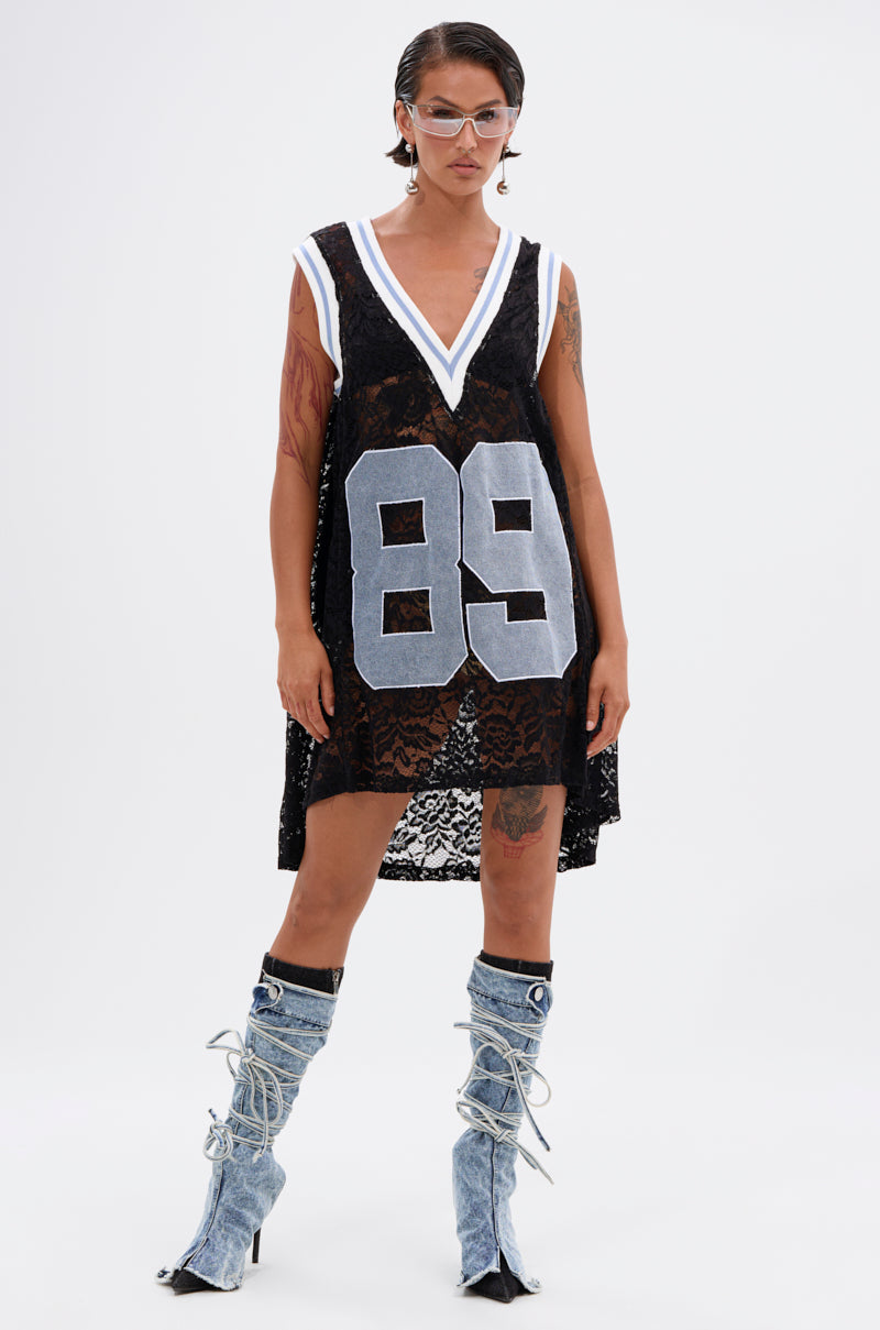Wearing the ON THE SIDELINES LACE JERSEY MINI DRESS WITH DENIM, a short-haired person with clear glasses and knee-high blue denim lace-up boots stands against a plain white background. The dress features a bold white "89" on the front.