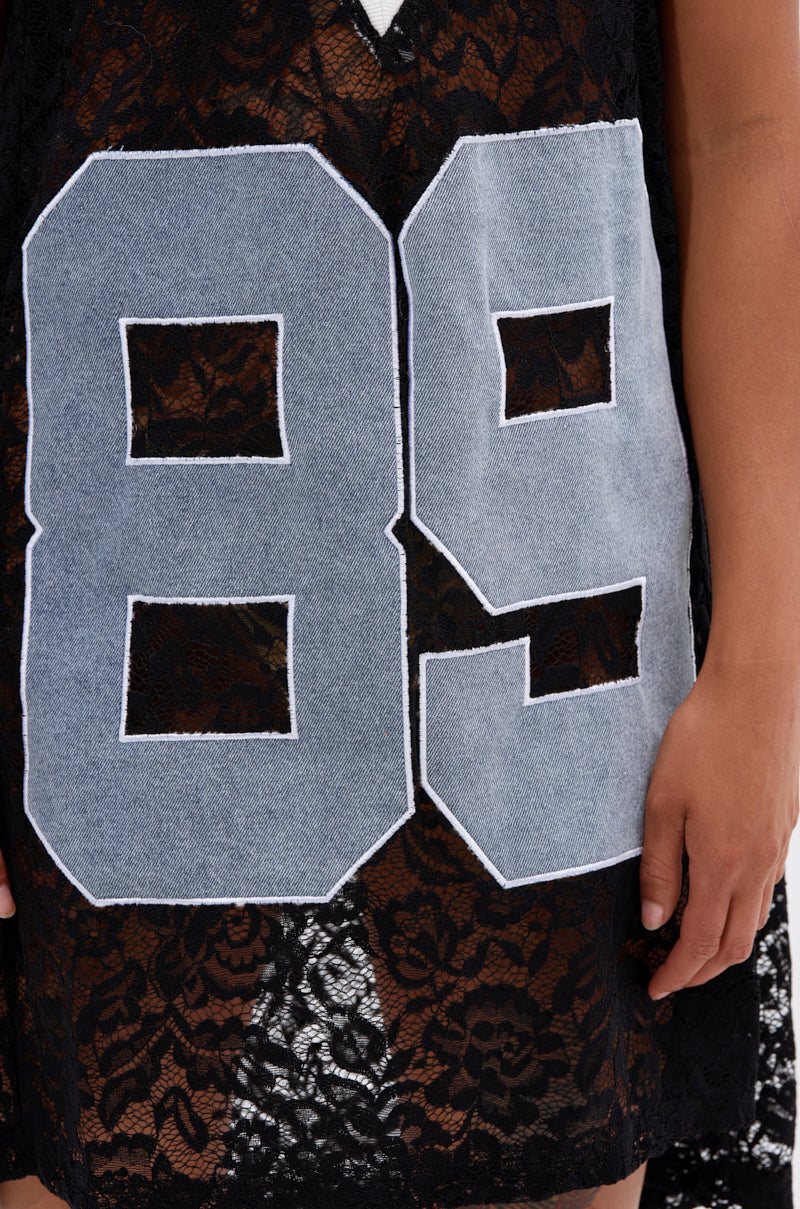 Close-up of the ON THE SIDELINES LACE JERSEY MINI DRESS WITH DENIM—a black lace mini dress with a bold light blue denim "89" stitched on the front. A hand appears at the right edge, highlighting the varsity style.