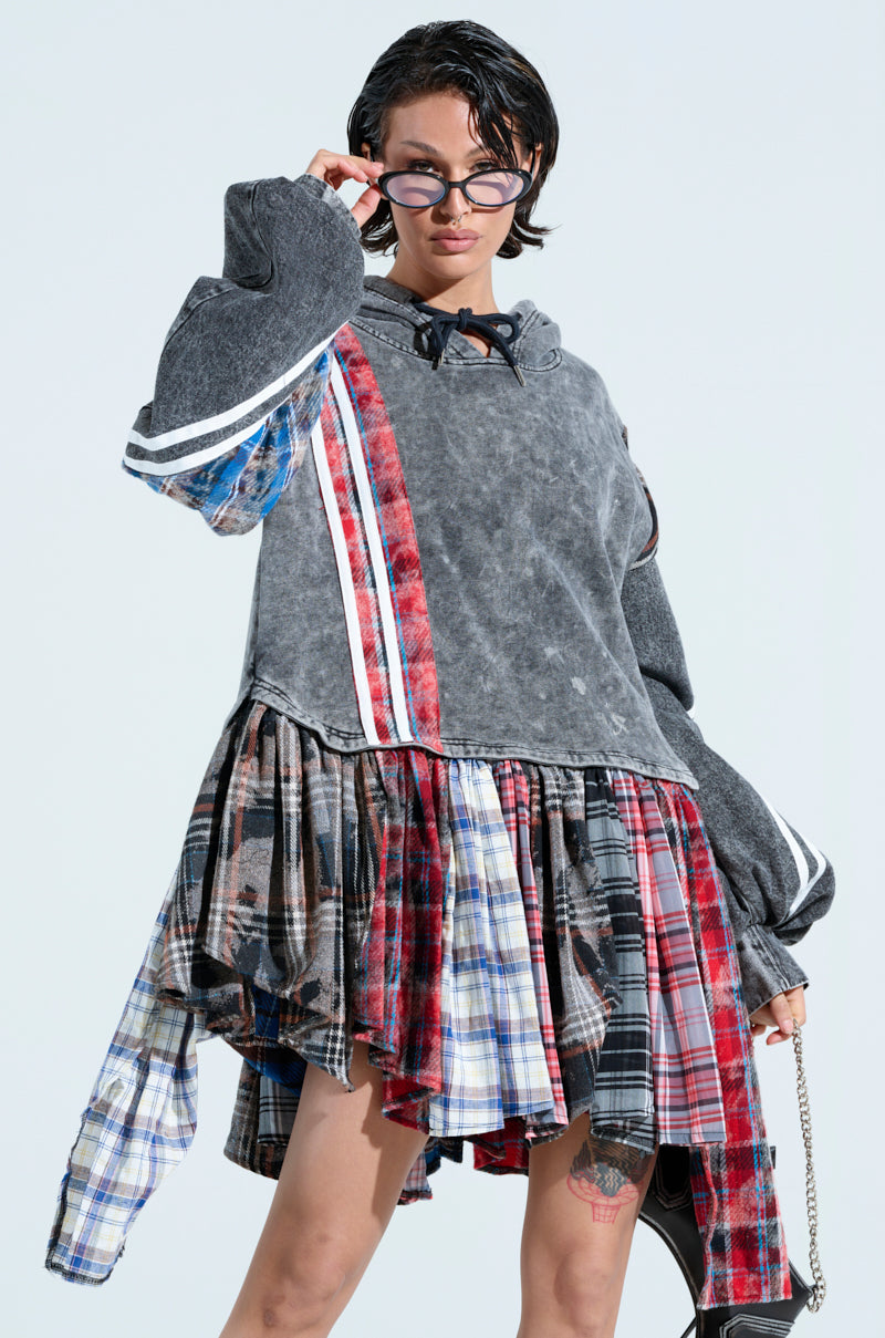 Wearing an oversized "A LITTLE BIT OF EVERYTHING PLAID SWEATSHIRT DRESS" with plaid patterns and a gray hood, the person poses against a light background, holding black-framed sunglasses near their face and carrying a chain-strap bag. Their hair is styled short and wet.