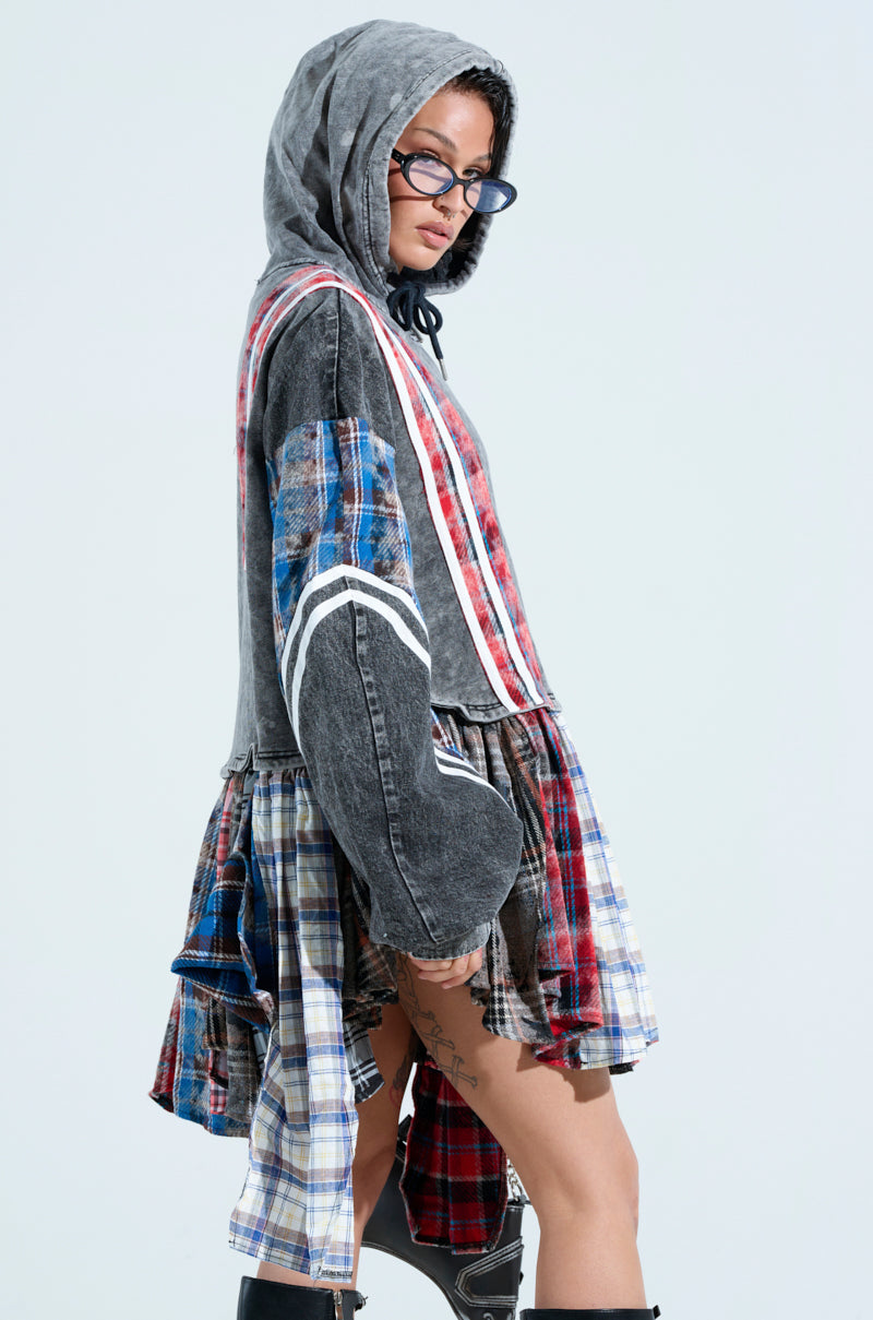A person is wearing "A Little Bit of Everything Plaid Sweatshirt Dress," featuring a mix of denim and plaid patterns. The cozy cotton jacket has loose sleeves, a flared hem, and offers an oversized fit. They have glasses, short hair, knee-high boots, and stand against a light background.