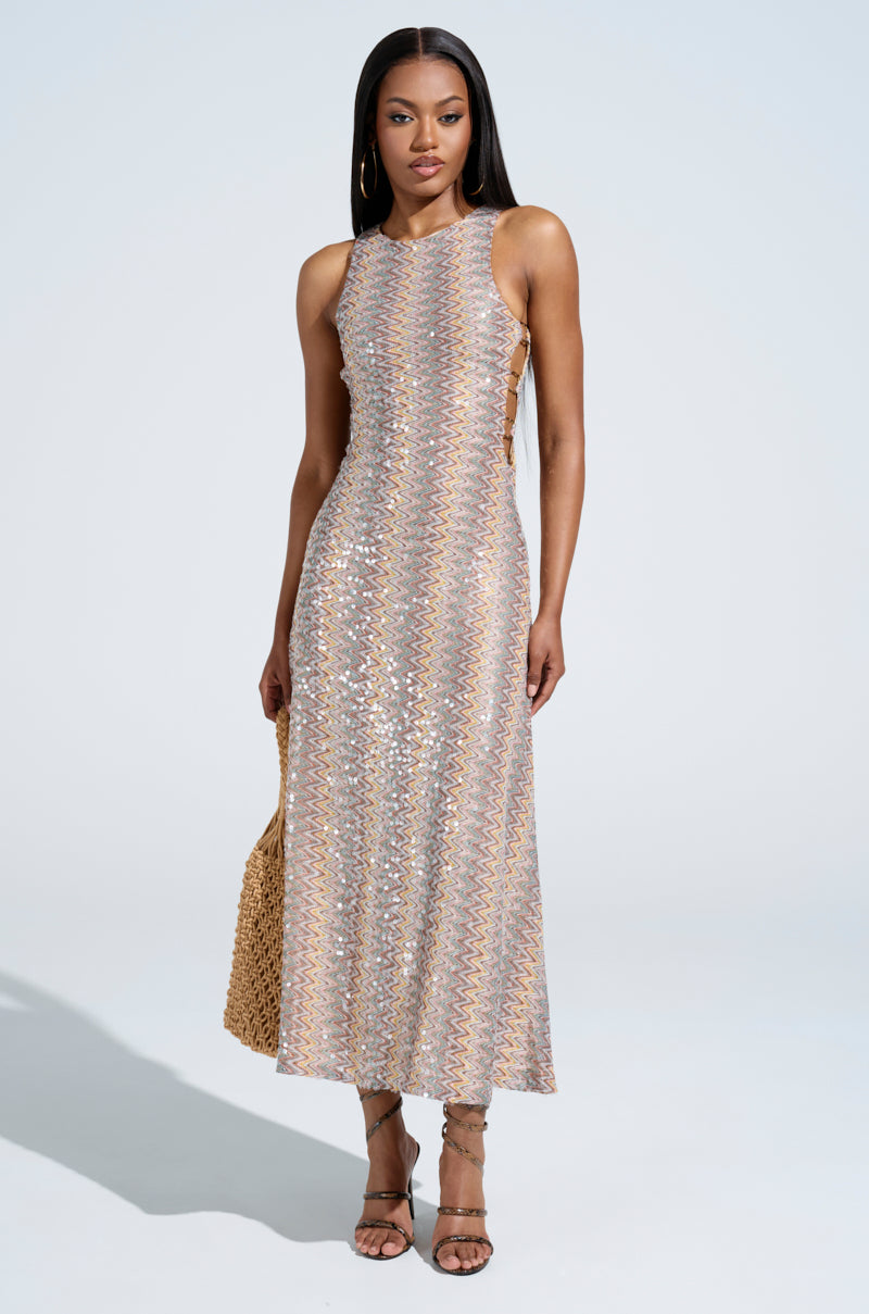 A woman exudes confidence in the "WOKE UP IN IBIZA CROCHET MIDI DRESS," a sleeveless, sparkling, multicolored sequined gown. She complements it with a woven bag and strappy heels, all set against a plain, light backdrop that highlights her chic ensemble.