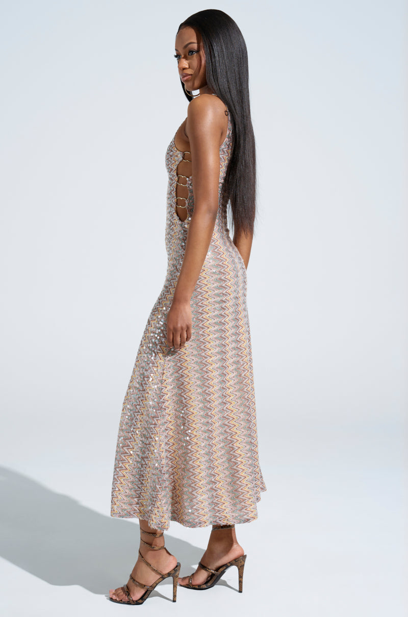 A person with long hair stands sideways wearing the WOKE UP IN IBIZA CROCHET MIDI DRESS, featuring a sleeveless, patterned crochet design and open back. They stand on a plain, light background in high heels.