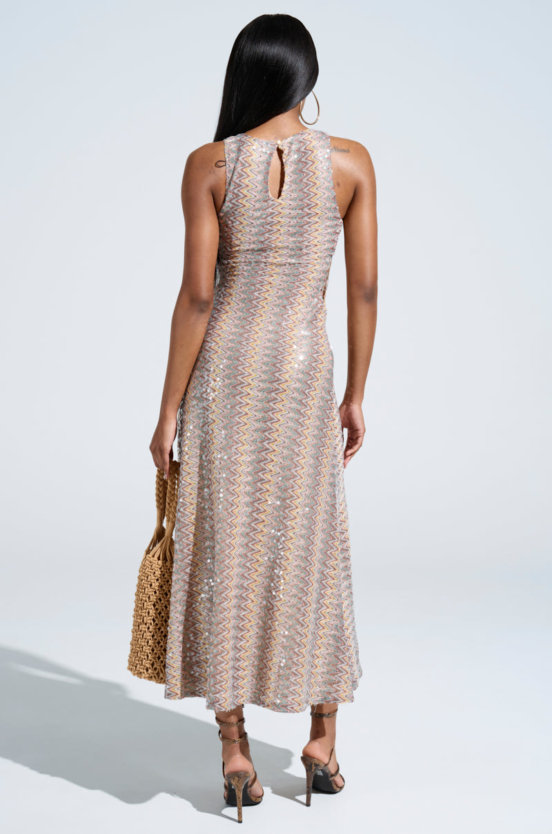 A person with long hair is seen from behind wearing the "Woke Up in Ibiza" crochet midi dress with a multicolored sequined pattern, holding a woven handbag and wearing strappy high heel sandals, set against a light background.