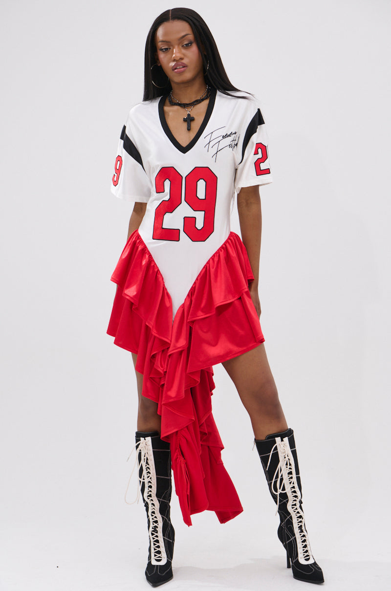 A woman models the FEMME FOOTBALL JERSEY MINI DRESS, a sporty V-neck dress with number 29 and red ruffled satin tiers, paired with black knee-high boots with white laces, against a plain white background.