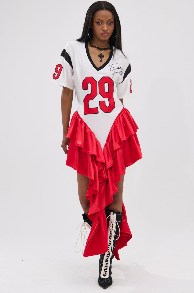 A woman models the FEMME FOOTBALL JERSEY MINI DRESS—a sporty, red and white V-neck dress with number 29, ruffled satin asymmetric skirt—paired with lace-up knee-high boots against a plain white background.