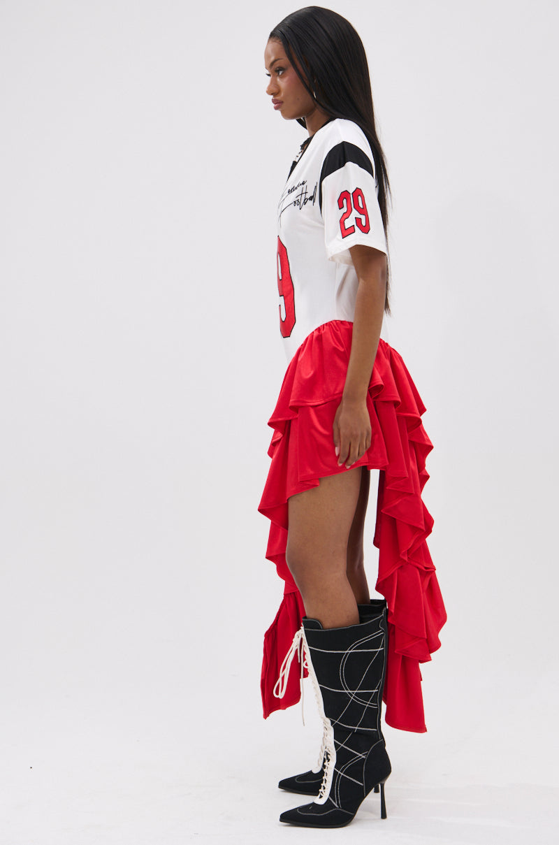 A woman in profile wears the FEMME FOOTBALL JERSEY MINI DRESS, a sporty V-neck dress with number 29 and bold red ruffled satin high-low skirt, paired with knee-high black lace-up stiletto boots.