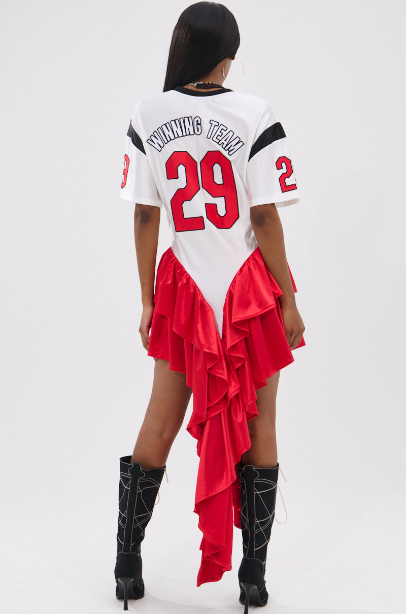 A person is wearing the FEMME FOOTBALL JERSEY MINI DRESS, a white V-neck dress with red number 29 and "WINNING TEAM" on the back, finished with asymmetrical red ruffled satin tiers, styled with black lace-up heeled boots.