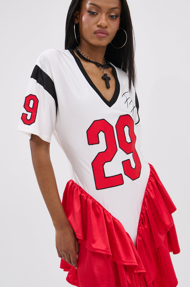 A woman models the FEMME FOOTBALL JERSEY MINI DRESS in white, featuring a sporty V-neck, red ruffled satin tiers, bold red 29 on the front, black sleeve stripe accents, and a cross pendant necklace.