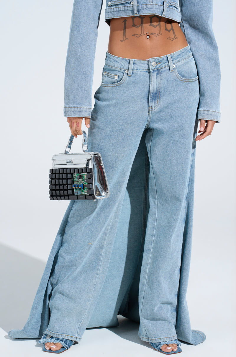 Person wearing SHAKE IT OFF DENIM PANT WITH SKIRT ATTACHED, featuring a light blue, wide-leg design and a matching cropped jacket. A small metallic handbag complements the tattooed midriff, while denim-strapped open-toe heels stand out against a plain white background.