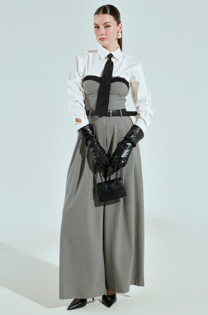 Against a plain backdrop, an individual exudes refined elegance in a white BOSS ENERGY BUTTON DOWN shirt and a long gray poplin dress with a black belt. They accessorize with black leather gloves, clutch a small black handbag, and wear pointed black shoes. Their hair is neatly pulled back.