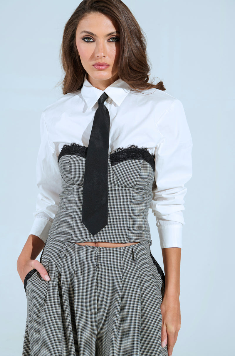 Wearing a Boss Energy Button Down, black tie, and houndstooth bustier and skirt, a woman stands confidently with one hand in her pocket. Her long brown hair complements her expression against a simple background, capturing the chic essence of the Akira Label style.