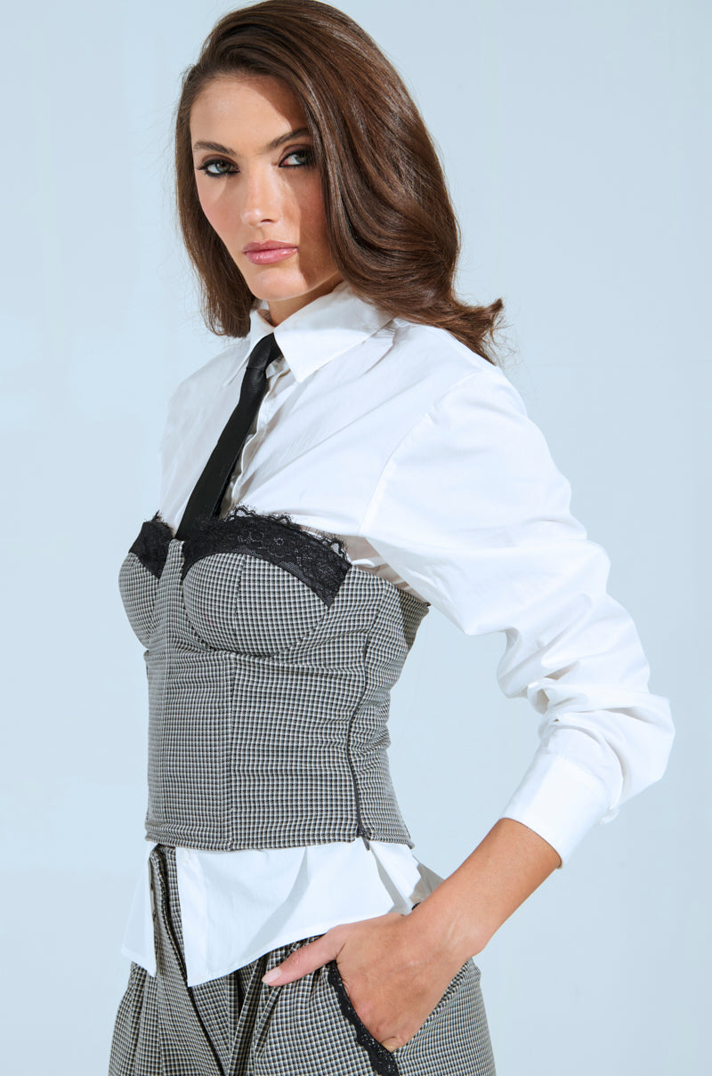 A woman with long brown hair wears a BOSS ENERGY BUTTON DOWN over a gray plaid corset top and matching pants from the AKIRA Label. She poses with her hands in her pockets against a minimalist backdrop.