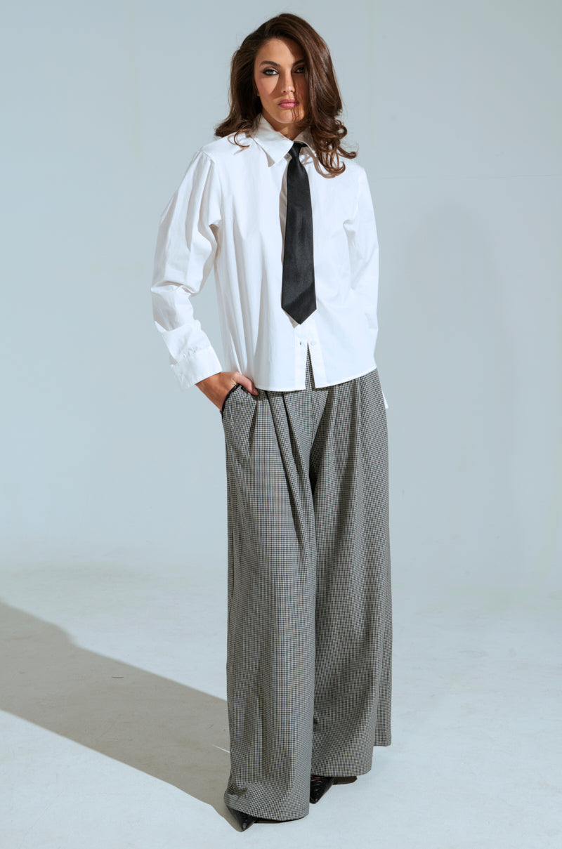 Confidently posed against a plain backdrop, a person wears the AKIRA Label BOSS ENERGY BUTTON DOWN with gray pinstripe pants. The loose white shirt, featuring a black tie, complements their long brown hair and relaxed hand-in-pocket pose.