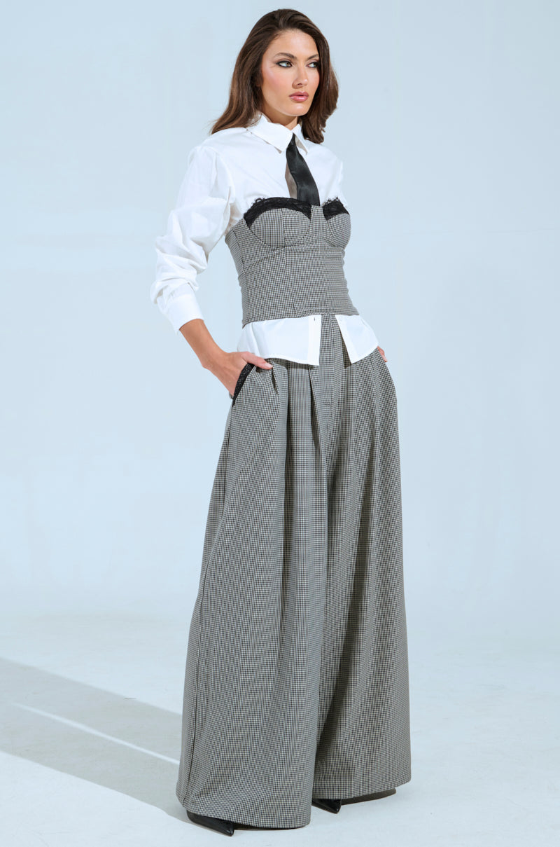 A woman poses against a neutral background in a fashionable AKIRA Label outfit, combining the white Boss Energy Button Down shirt with a tie, topped with a gray strapless corset and wide-legged gray trousers. Her hair flows effortlessly as she casually slides one hand into her pocket.