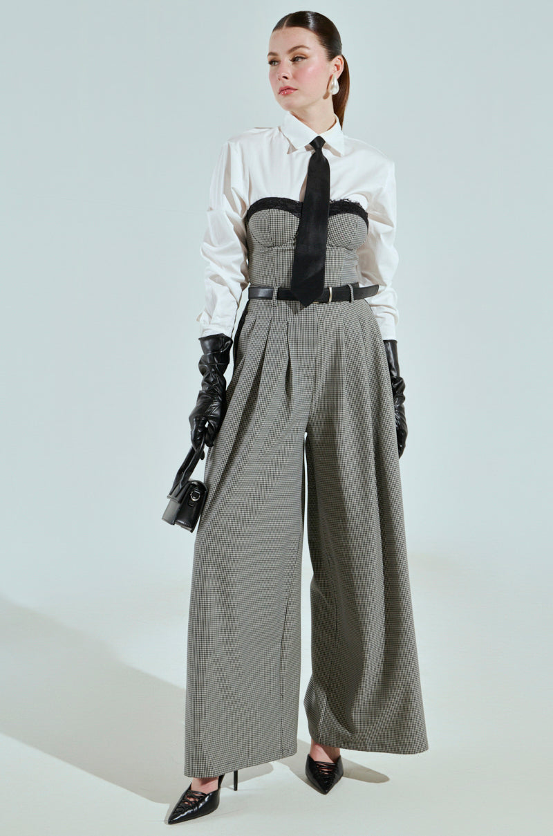Wearing the BOSS ENERGY BUTTON DOWN with a black tie, AKIRA Label corset-like top, wide gray trousers, black leather gloves, and pointed black shoes, they exhibit confidence. They hold a small handbag against a minimalist white background.