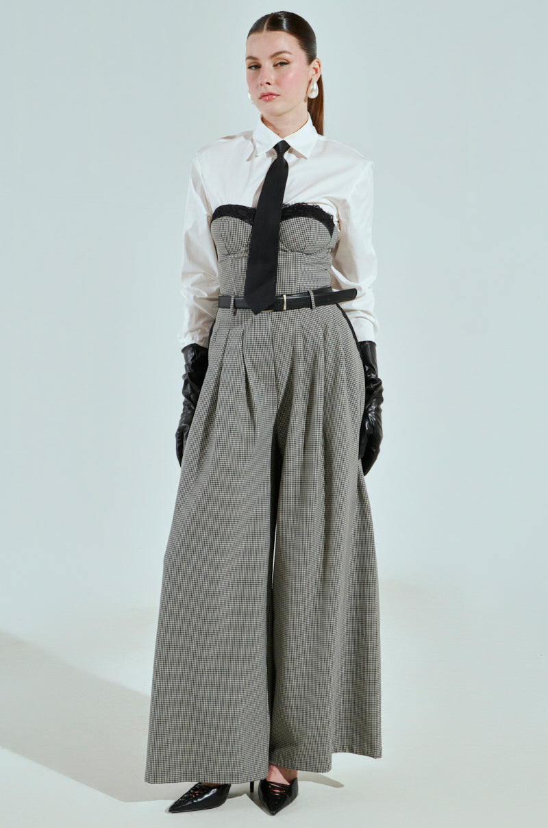 A person exudes Boss Energy in a white BOSS ENERGY BUTTON DOWN shirt, black tie, and large gray pinstriped trousers. Black gloves and pointed shoes complete the outfit, with long hair pulled back against a plain background.