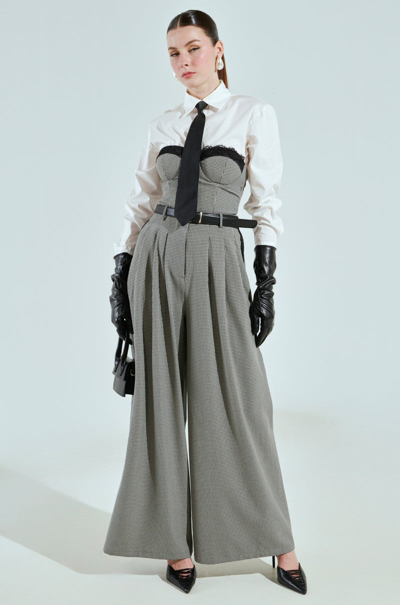 A person poses confidently in high-waisted wide-leg trousers and a BOSS ENERGY BUTTON DOWN, layered with a corset belt. Accessories include leather gloves, high heels, a black tie, sleek black purse, and large earrings. With hair pulled back, they exude pure elegance.