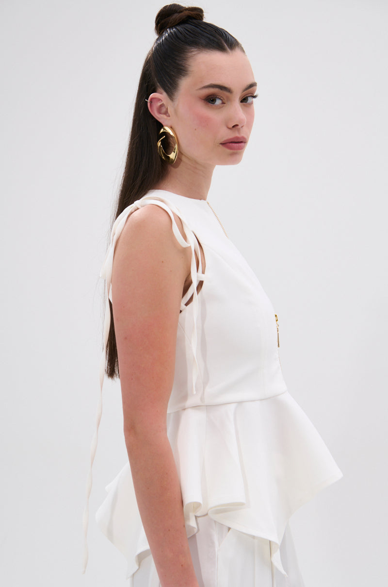 A woman with long brown hair in a topknot wears the PEPLUM FRONT ZIP SLEEVELESS BLOUSE IN WHITE and large gold hoop earrings, standing against a plain light background.