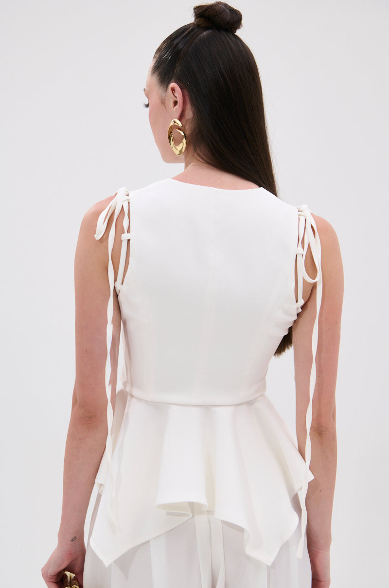 A woman with long, straight dark hair faces away from the camera, wearing the PEPLUM FRONT ZIP SLEEVELESS BLOUSE IN WHITE. The blouse features shoulder tie details and a flared hem, showcasing its elegant back design.