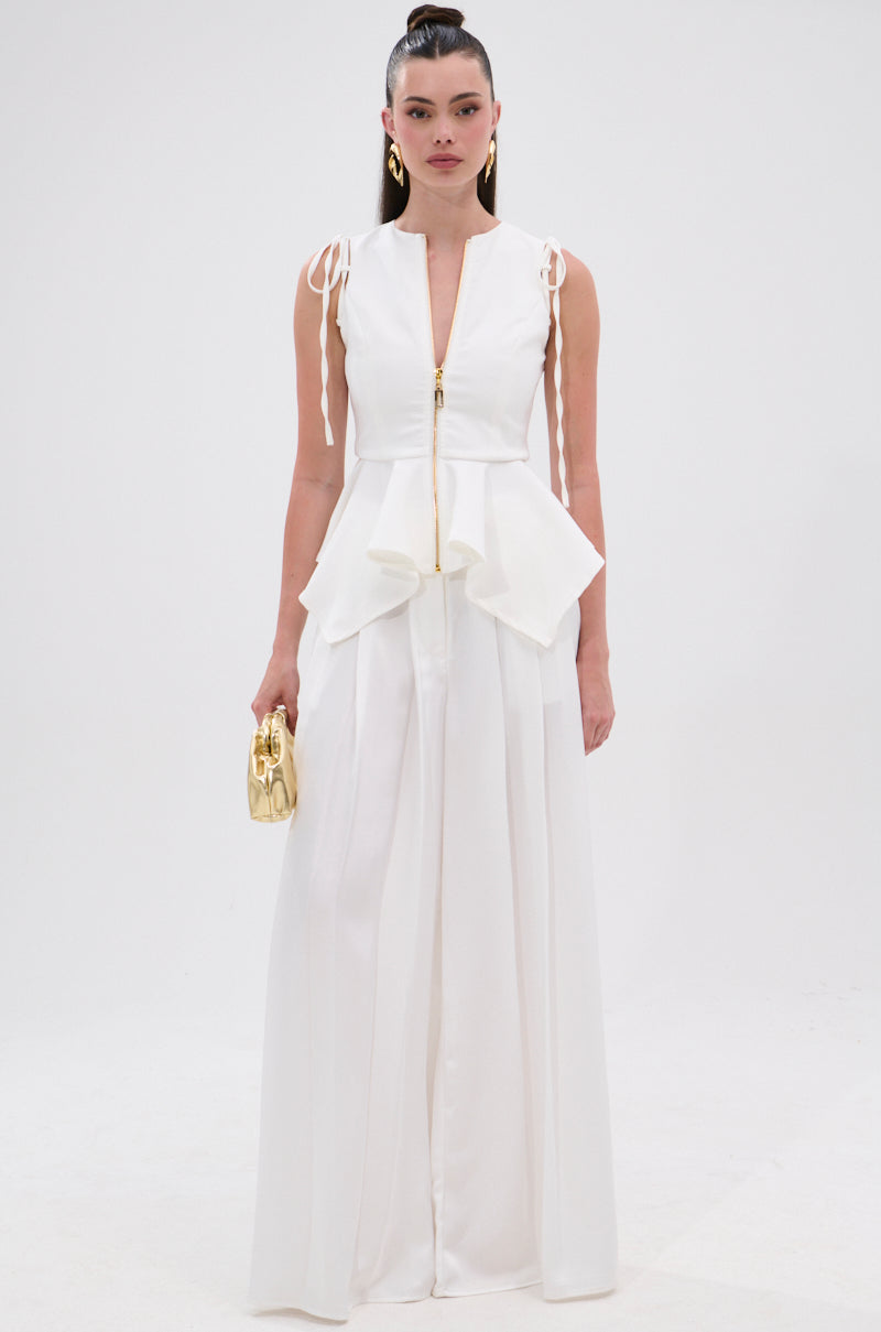 A woman stands against a white background wearing the PEPLUM FRONT ZIP SLEEVELESS BLOUSE IN WHITE with a pleated skirt, holding a small metallic gold handbag and her hair in a high ponytail.