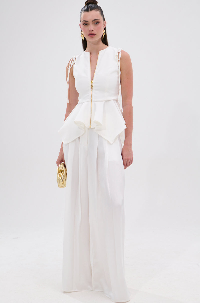 A woman stands against a white background wearing the PEPLUM FRONT ZIP SLEEVELESS BLOUSE IN WHITE, styled with a deep V-neck and peplum detail. She accessorizes with a gold clutch, gold hoop earrings, and slicked-back hair.