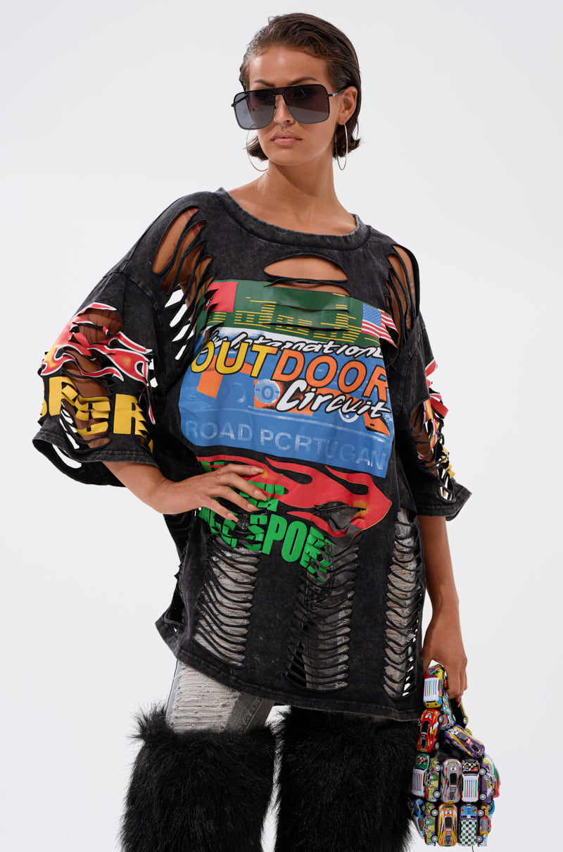 A woman in oversized sunglasses, the OFF TO THE RACES DISTRESSED OVERSIZED TEE, metallic leggings, furry black leg warmers, and holding a small multicolored bag stands against a plain white background.