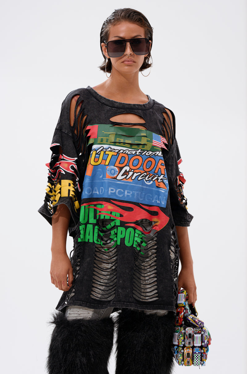 Wearing oversized sunglasses, furry black pants, and holding a multicolored patterned bag, a person models the OFF TO THE RACES DISTRESSED OVERSIZED TEE with racing-inspired graphics against a plain white background.