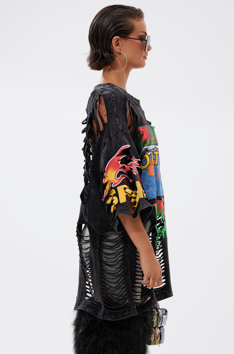 A woman in large sunglasses and hoop earrings wears the OFF TO THE RACES DISTRESSED OVERSIZED TEE with colorful prints and cut-out details, paired with textured black pants, standing in profile against a plain background.