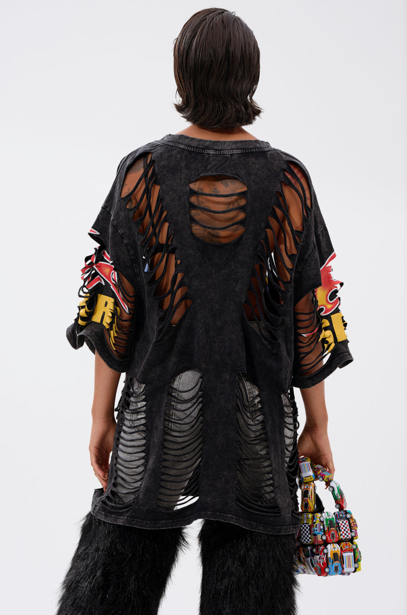 A person with short dark hair, seen from behind, wears the OFF TO THE RACES DISTRESSED OVERSIZED TEE with large rips, black furry pants, and holds a colorful beaded bag against a plain white background.