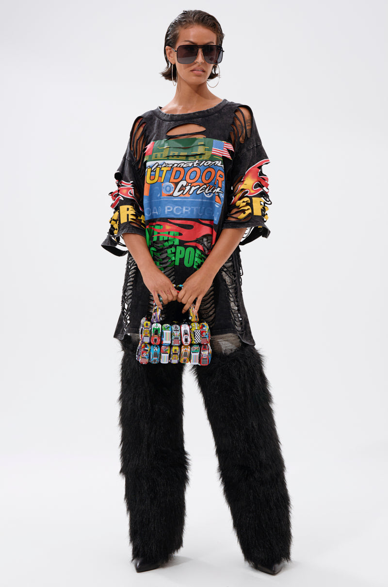 A person in the OFF TO THE RACES DISTRESSED OVERSIZED TEE, black furry pants, oversized sunglasses, and holding a small handbag adorned with toy cars stands against a plain white background.