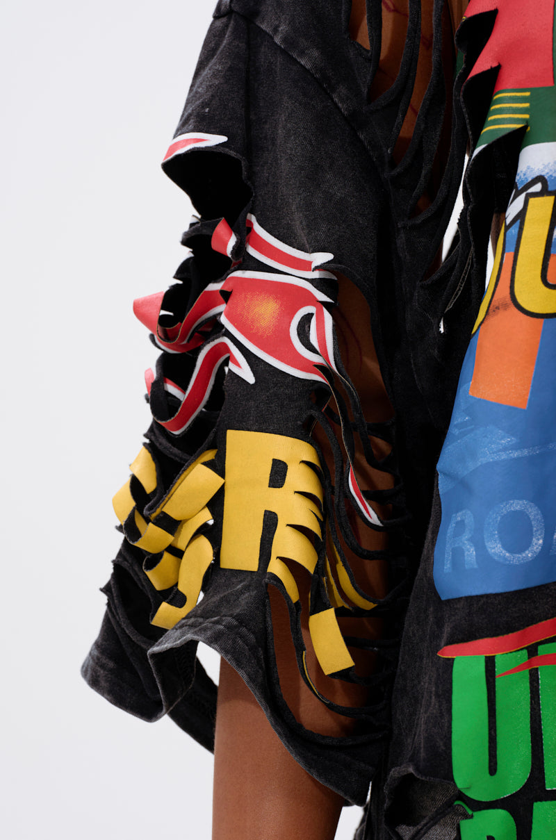 A close-up of a person's arm wearing the OFF TO THE RACES DISTRESSED OVERSIZED TEE, featuring colorful racing-inspired graphics and multiple horizontal slashes that reveal skin. The background is plain and light.