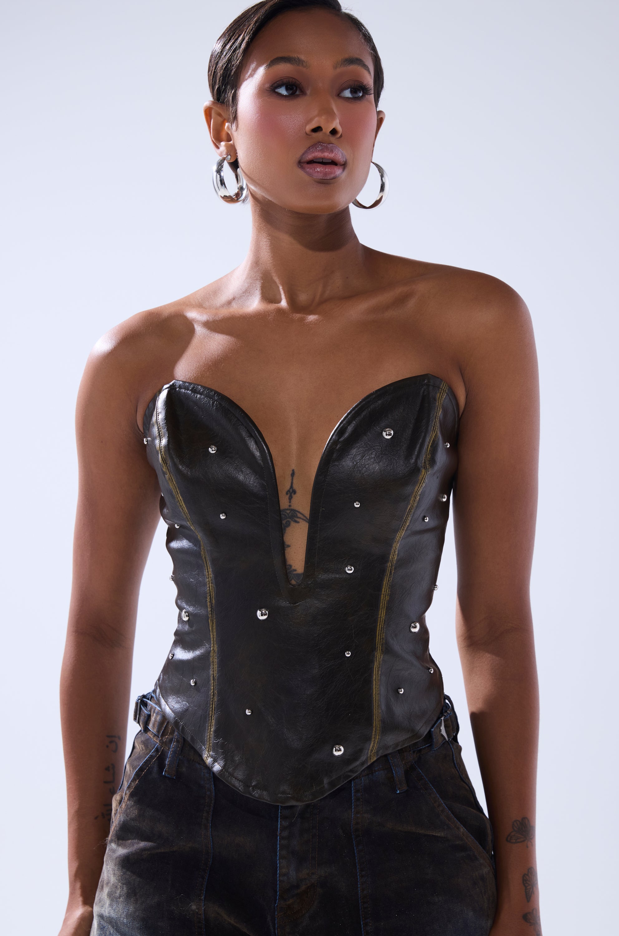 A woman with short hair, hoop earrings, and visible tattoos wears the HARD TO PLEASE STUDDED FAUX LEATHER CORSET with dark pants, standing against a plain light background.