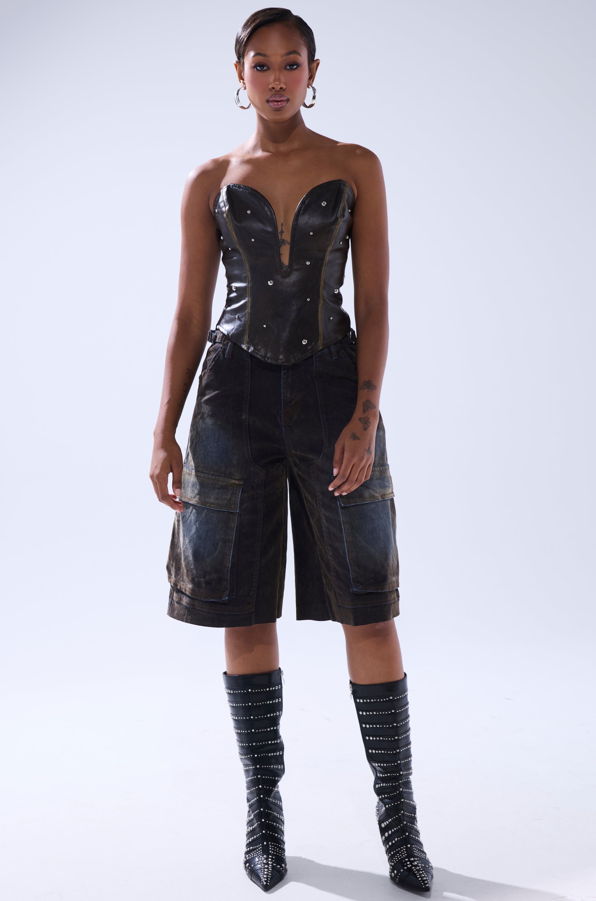 A woman faces forward in the HARD TO PLEASE STUDDED FAUX LEATHER CORSET, loose knee-length shorts, and knee-high black boots with silver studs. She has short hair, hoop earrings, tattoos on her left arm, and a plain white background.