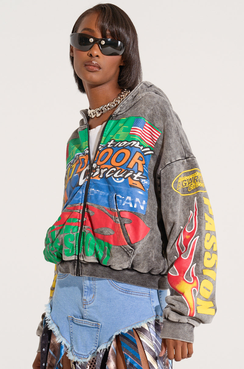 A person wearing black sunglasses, an OFF TO THE RACES GRAPHIC ZIP UP HOODIE featuring racing-inspired logos and text in a mineral wash fabric, and a denim mini skirt with a frayed hem. The background is plain white, making the vibrant colors of the hoodie and outfit stand out.