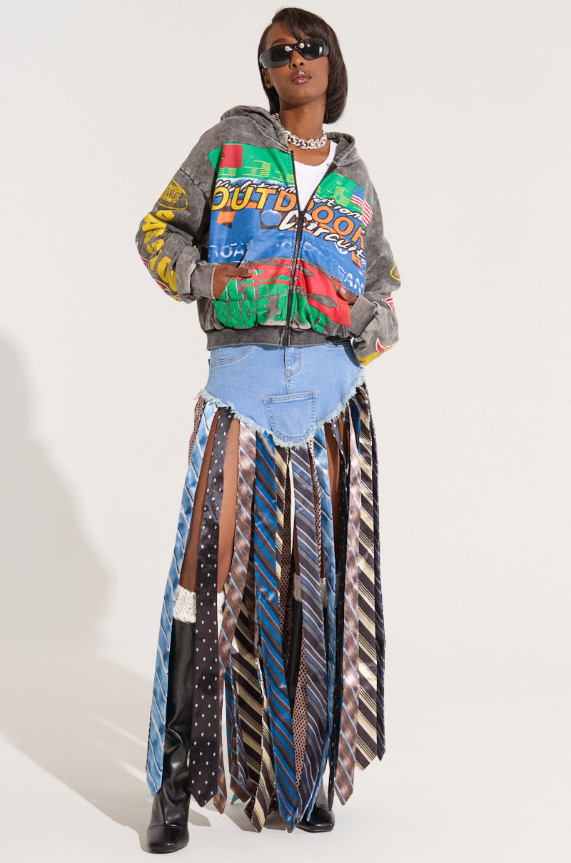 A person wearing sunglasses stands confidently with their hands in their pockets. They have on an "OFF TO THE RACES" graphic zip-up hoodie, a denim skirt, and a unique skirt extension made from various neckties. The background is plain white.