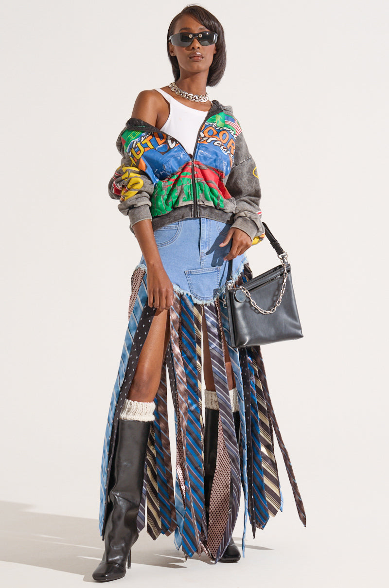 A stylish person poses against a plain background, wearing the colorful "Off to the Races" graphic zip up hoodie over one shoulder, a white top, a unique skirt made of blue ties, and knee-high black boots. Accessories include a silver chain necklace, black sunglasses, and a black leather handbag.