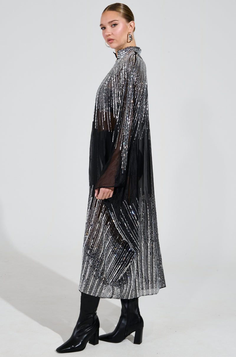 A person wearing a glamorous SPARKLE AND SHINE BLOUSE, embellished with silver shiny stripes and mirrored sequins, stands facing sideways. The blouse features sheer sections made from sheer nylon fabrication and is paired with tall black boots against a plain, light-colored background.