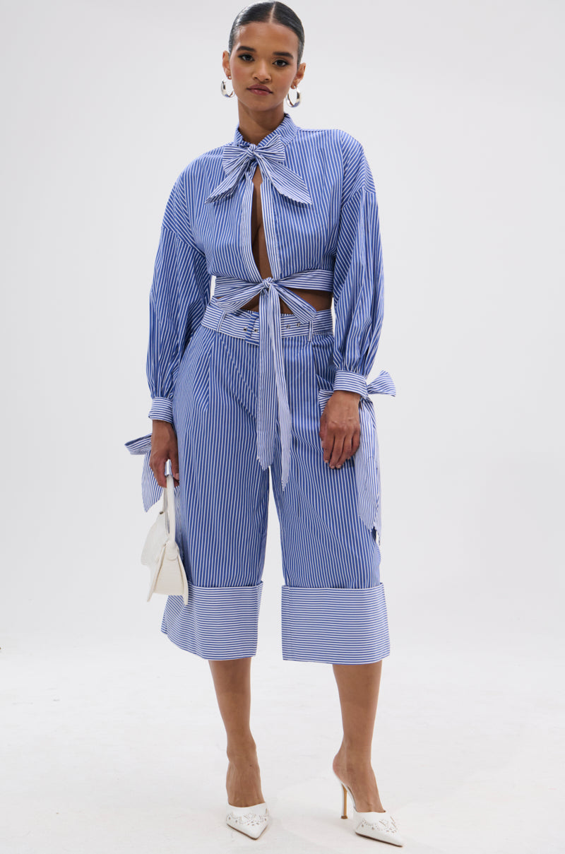 A woman models the UNDER THE SUN STRIPE TIE FRONT BLOUSE—a blue-and-white striped, cropped, boxy top—paired with matching wide-leg capri pants, a white handbag, and white pointed-toe heels.