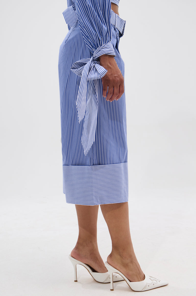 A person is standing on a white background, wearing UNDER THE SUN STRIPE SHORTS, a blue and white vertical striped dress with tied sleeves, and white high-heeled shoes.
