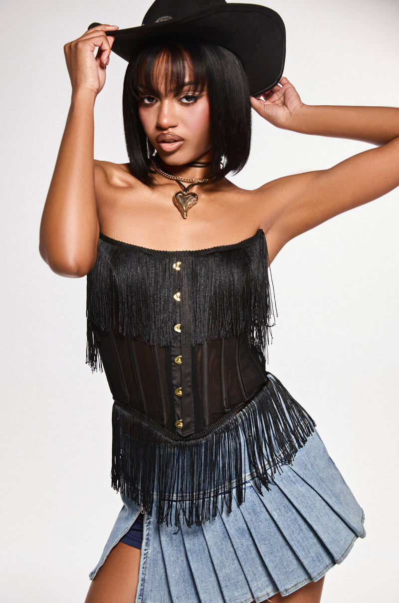 Wearing the FLAPPER GIRL CORSET TOP, a woman exudes vintage-chic flair with a black fringed style, paired with a blue denim pleated skirt and black cowboy hat. She confidently accessorizes with a choker necklace featuring a heart pendant, set against a neutral backdrop.