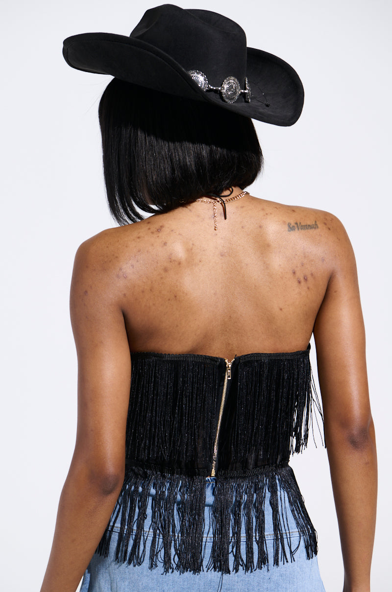 Back view of a person in a vintage-chic black strapless FLAPPER GIRL CORSET TOP with fringe, light blue jeans, and a black wide-brimmed hat with silver accents. Their back showcases natural freckles and a tattoo below the neck.