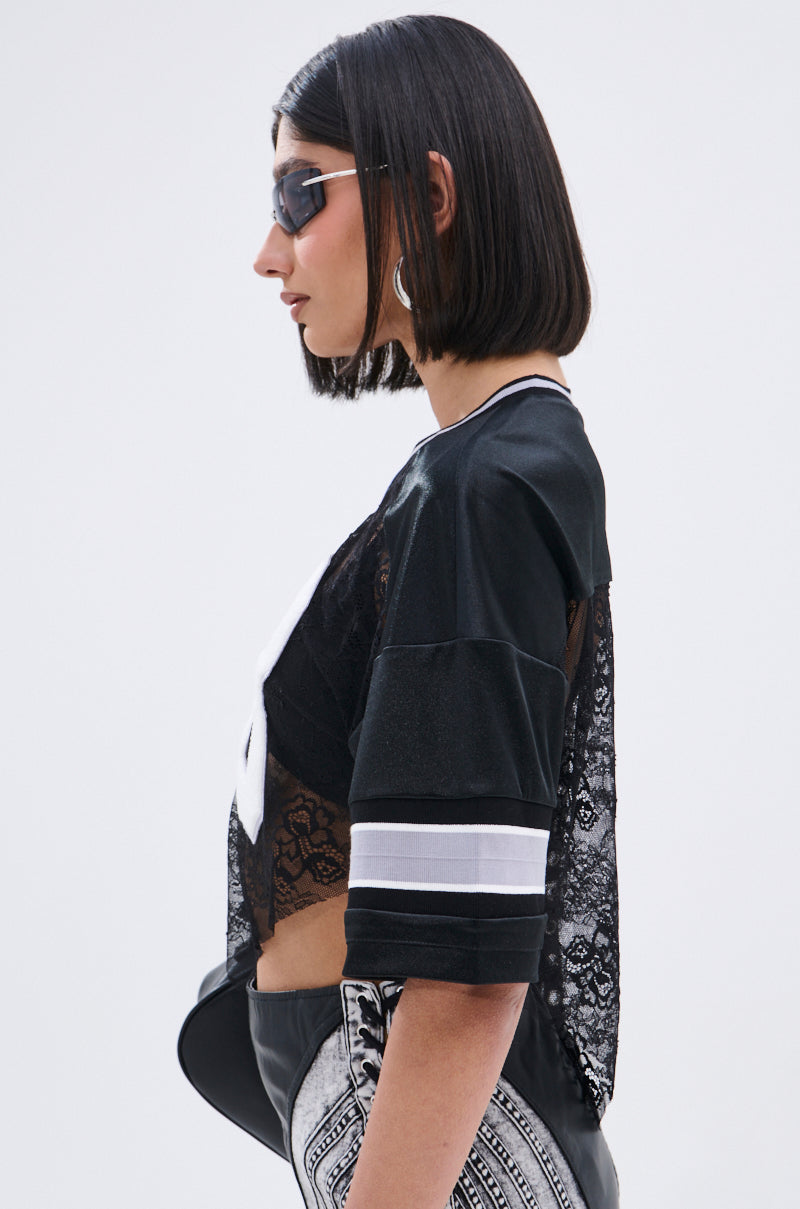 A woman in profile with straight, shoulder-length dark hair wears sunglasses, silver hoop earrings, and the LUCKY LACE JERSEY TOP—a sheer black lace top with white striped sleeves—against a plain light background.