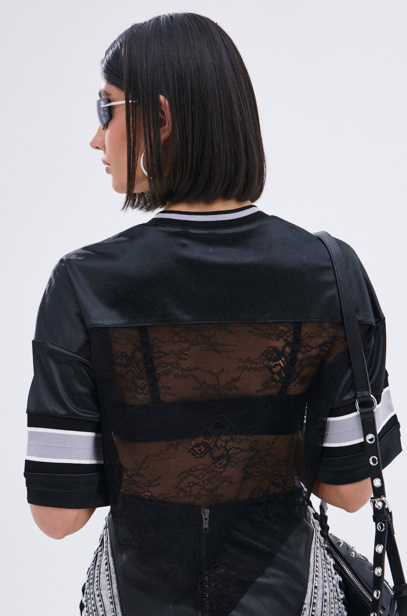 Wearing sunglasses and carrying a black studded bag, a person with straight, shoulder-length dark hair sports the LUCKY LACE JERSEY TOP, which features sheer black lace panels on the back and white striped sleeves.