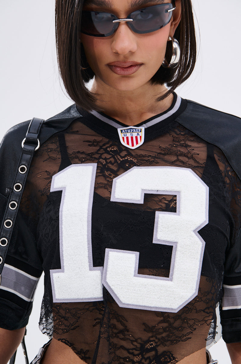 A woman wears the LUCKY LACE JERSEY TOP, a sheer black lace top with an oversized varsity number 13 patch, paired with sunglasses and hoop earrings.