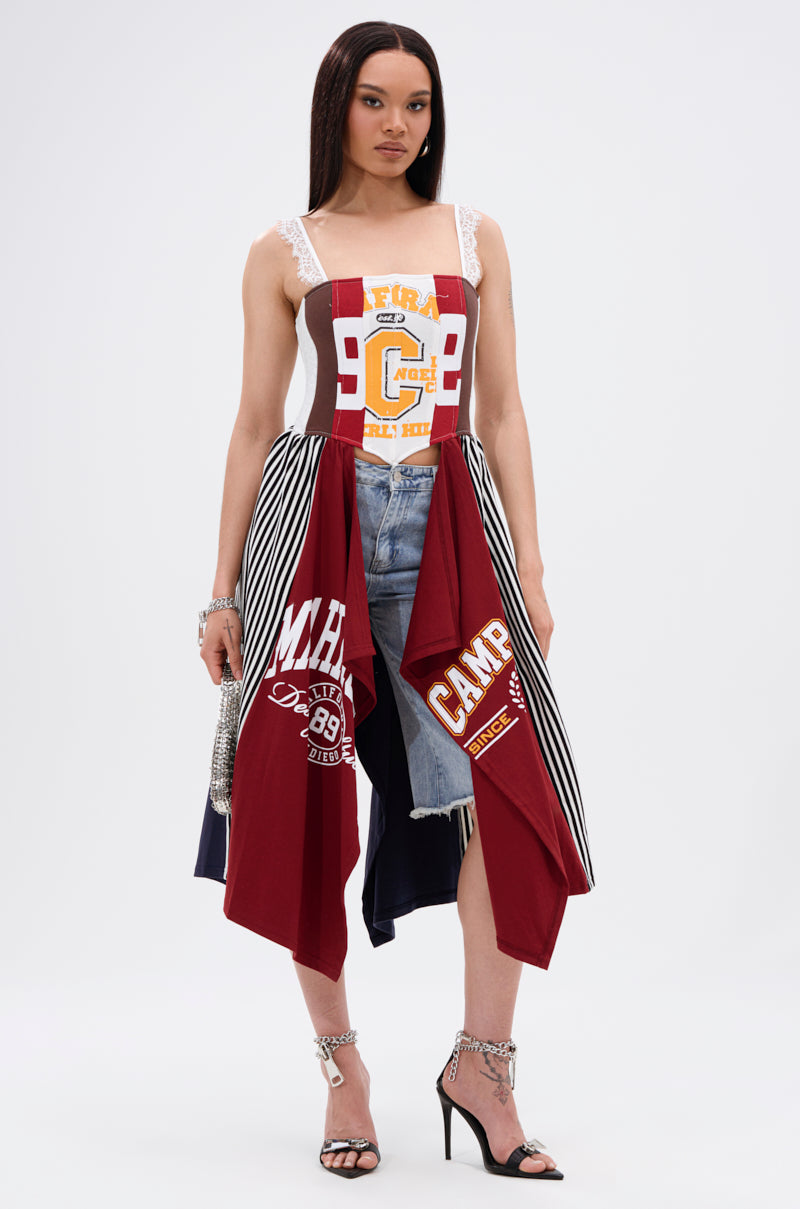A woman with straight hair wears the LIGHTS OUT PATCHED UP JERSEY CORSET, crafted from upcycled collegiate tees and striped fabric, layered over denim shorts and paired with high-heeled sandals against a plain background.