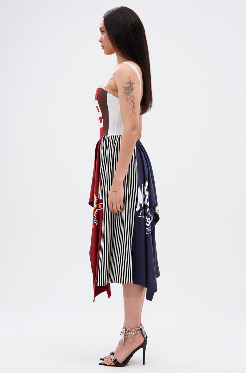 A woman stands in profile wearing the LIGHTS OUT PATCHED UP JERSEY CORSET, crafted from upcycled collegiate tees with a red, white, and blue patchwork design. She pairs it with high-heeled sandals, long straight hair, and a visible arm tattoo.