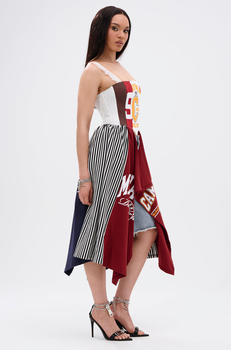 A woman stands in profile against a white background wearing the LIGHTS OUT PATCHED UP JERSEY CORSET, a sleeveless piece crafted from upcycled collegiate tees with striped, maroon, and graphic panels, paired with strappy high-heeled sandals.