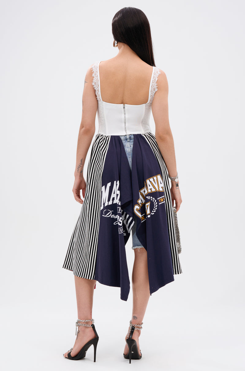 A woman faces away, wearing the LIGHTS OUT PATCHED UP JERSEY CORSET—a white sleeveless top with lace straps—paired with a unique skirt made from upcycled collegiate tees, high heels, and a small handbag.