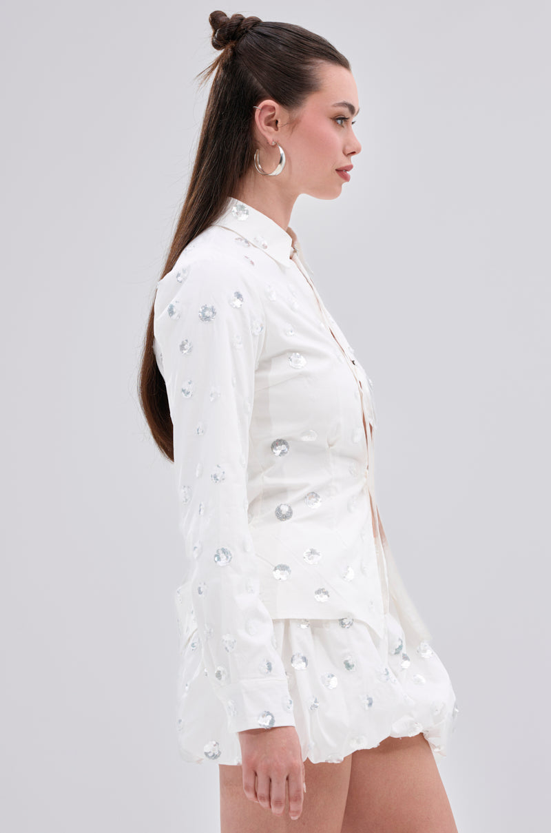 A woman poses in profile wearing the LUX FASHION BLOUSE, a white cotton-blend, long-sleeved top with shiny silver floral sequin embellishments and a flared skirt. Her long hair is styled half-up, half-down.