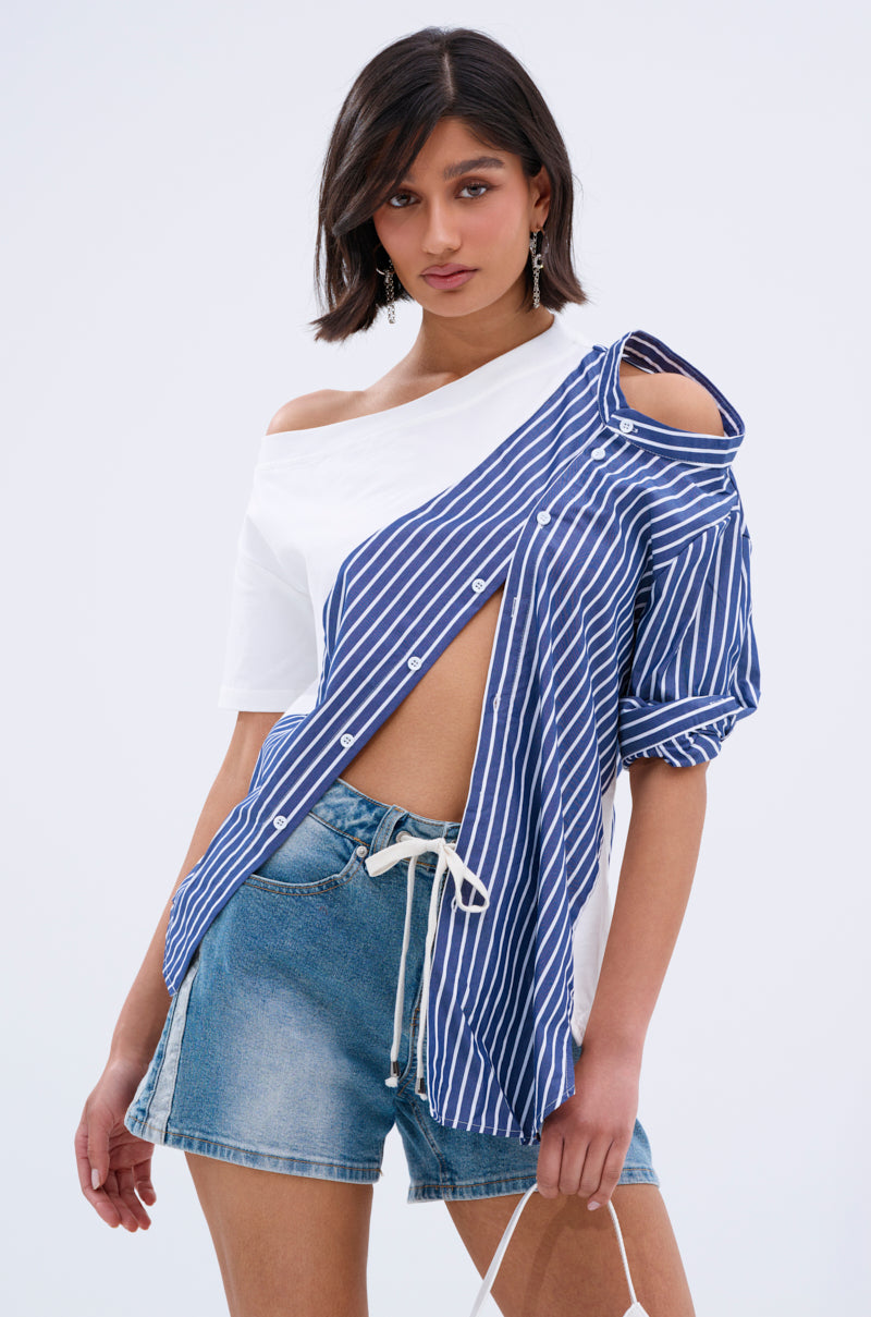 A woman with short dark hair wears the MIXED UP TSHIRT BLOUSE, featuring an asymmetrical design with one side white and the other blue-and-white stripes, styled off the shoulder and partially unbuttoned, paired with denim shorts.